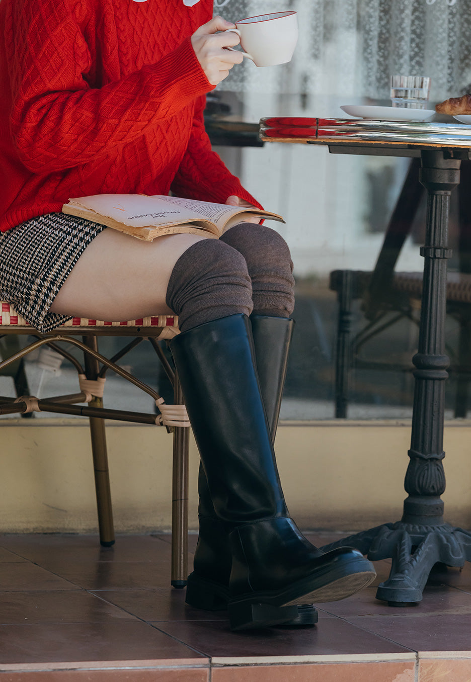 Person in a red sweater and green boots sitting at a table with a book and cup.