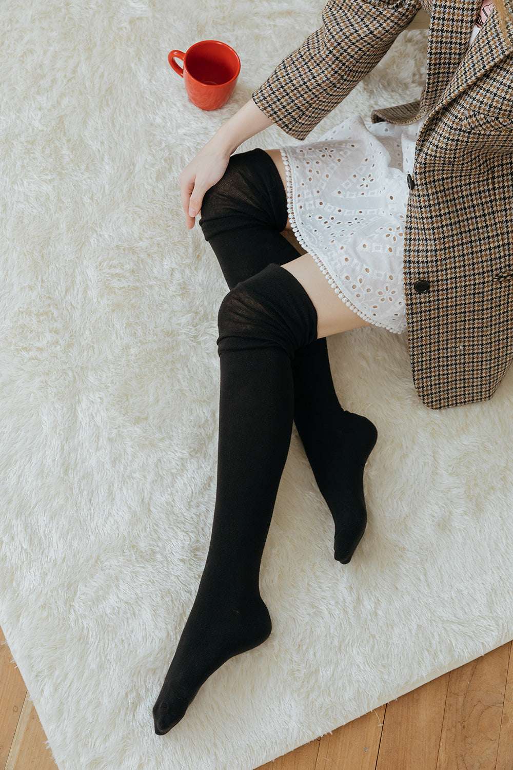 Person wearing black knee-high socks on a white rug with a red mug and plaid coat.