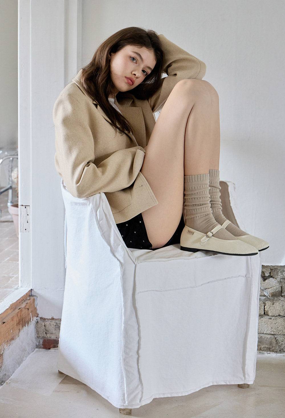 Woman sitting on a white chair wearing a beige coat, black shoes, and beige knee-high boots.