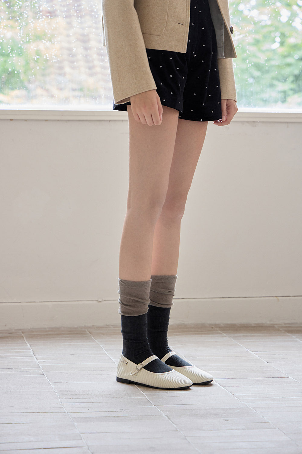 Person wearing a beige blazer, black skirt, and gray knee-high socks with black shoes in a room with large windows.