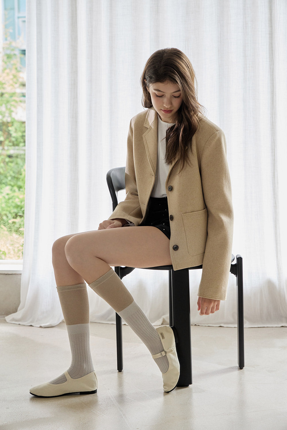 Woman sitting on a chair wearing a beige blazer, white shirt, black shorts, and white shoes.