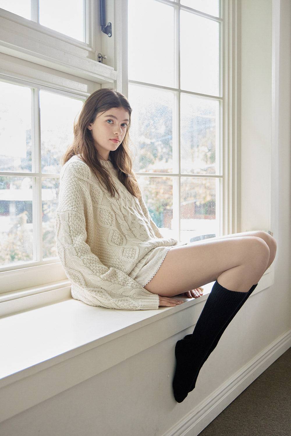 Woman sitting on a windowsill wearing a cream sweater and black stockings.