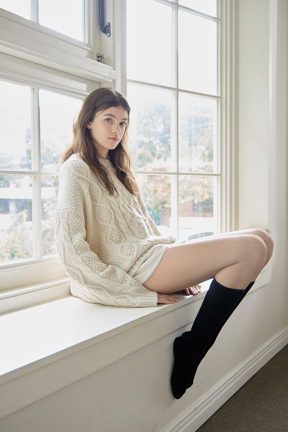 Woman sitting on a windowsill wearing a cream sweater and black stockings.