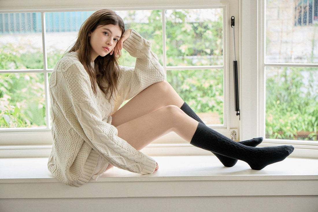 Woman sitting on a windowsill wearing a white sweater and black knee-high socks.