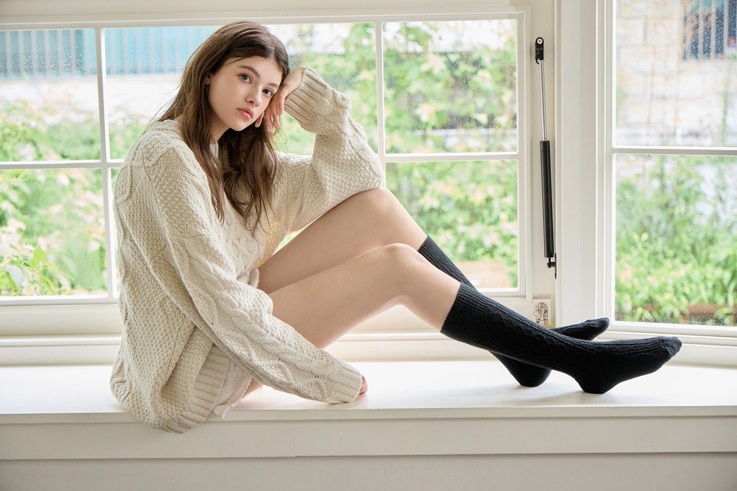 Woman sitting on a windowsill wearing a white sweater and black knee-high socks.