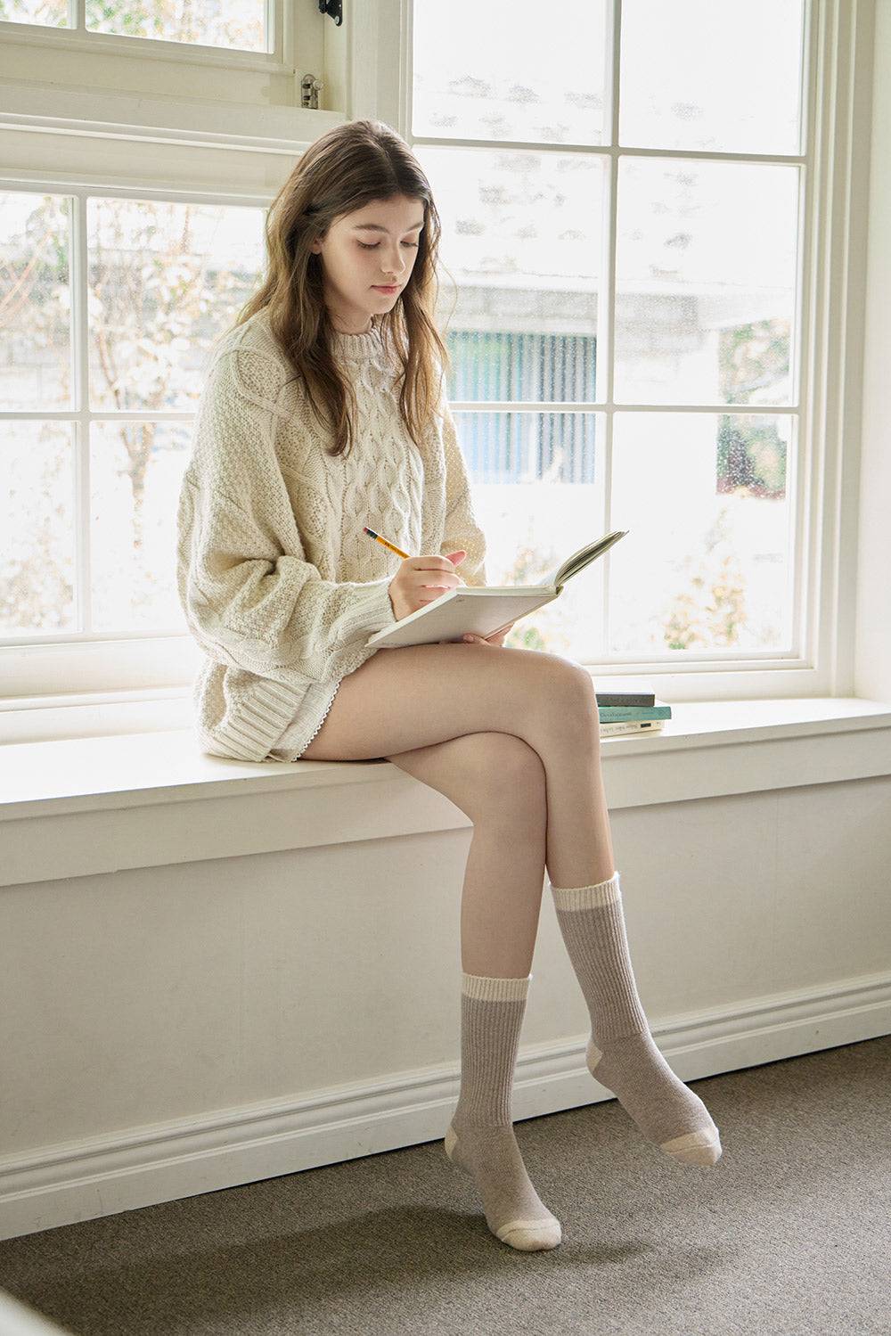 Woman sitting on a windowsill writing in a notebook with a pencil.