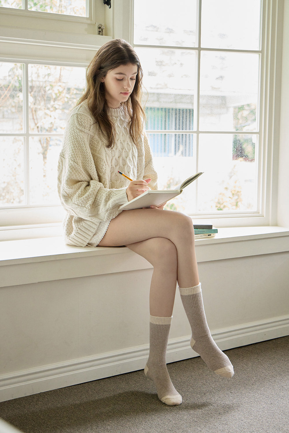 Woman sitting on a windowsill writing in a notebook with a pencil.