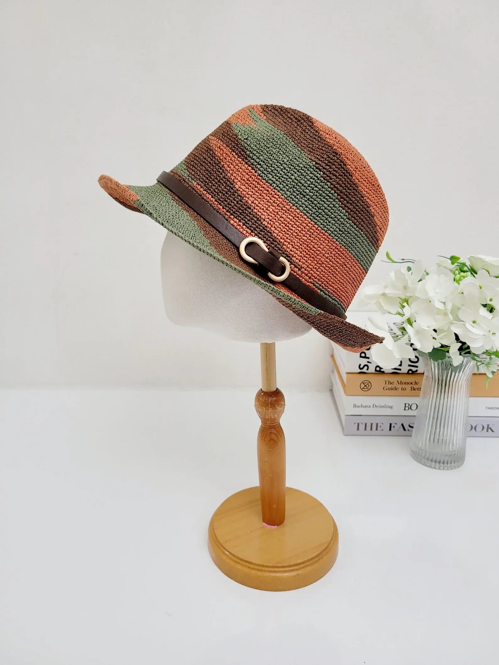 serengeti fedora hat with green and brown horizontal stripes, accented by a brown leather band and gold buckle, displayed on a wooden stand.