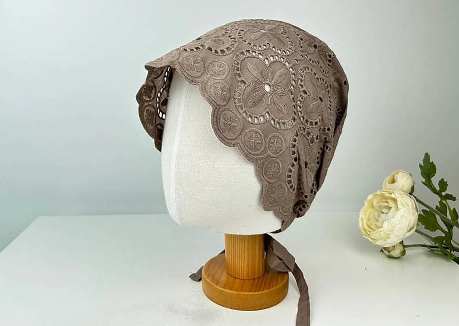 Taupe eyelet beanie with floral lace pattern, shown on a mannequin head with ribbon ties and a scalloped edge.