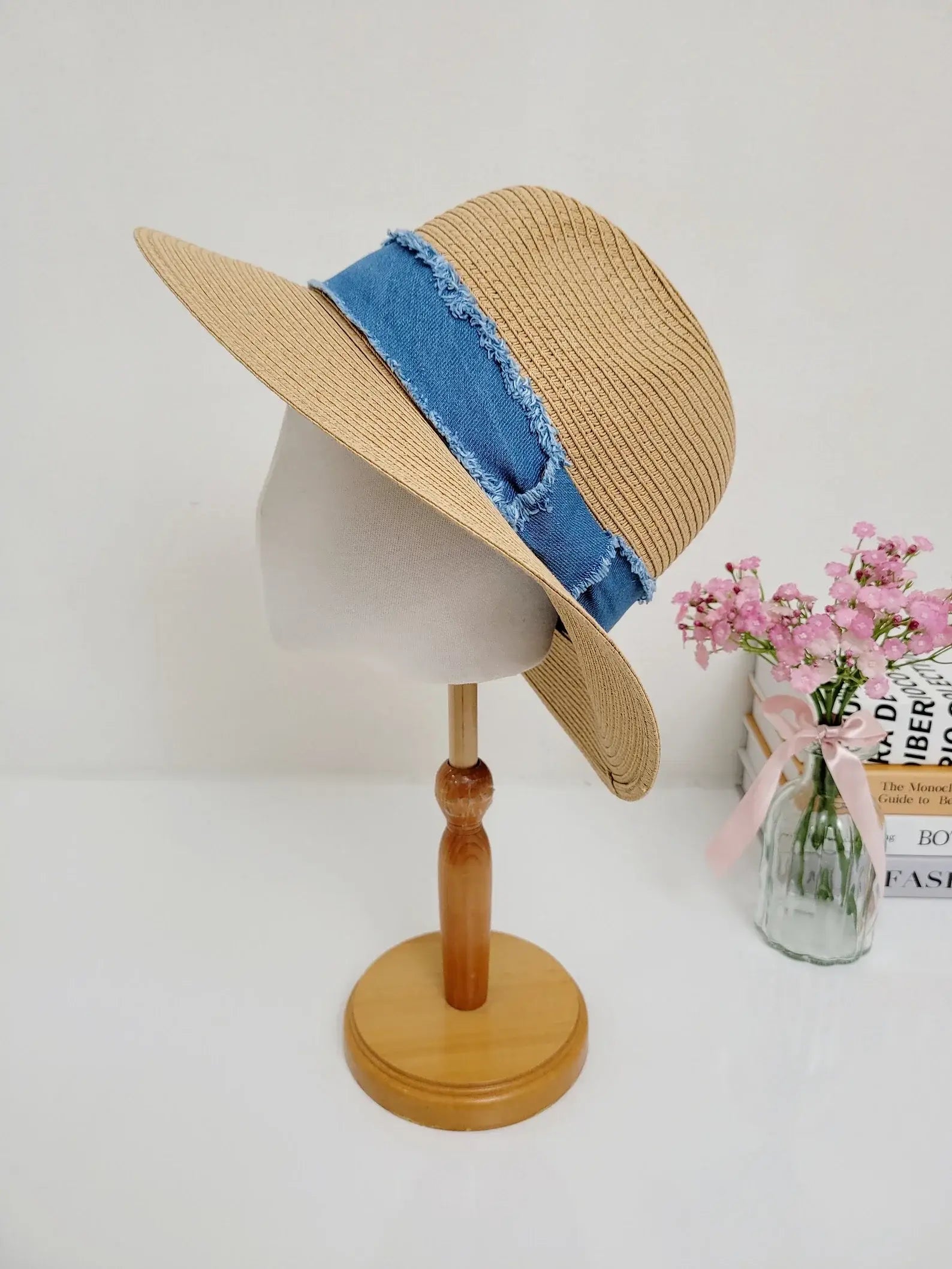 long island straw hat with a blue denim band on a wooden stand, next to a vase of pink flowers