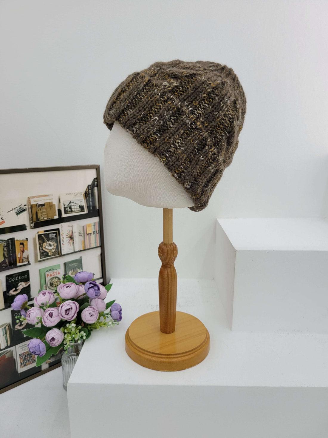 Knitted beanie on a lampshade with a wooden arm, next to a box of stamps and a bouquet of flowers.