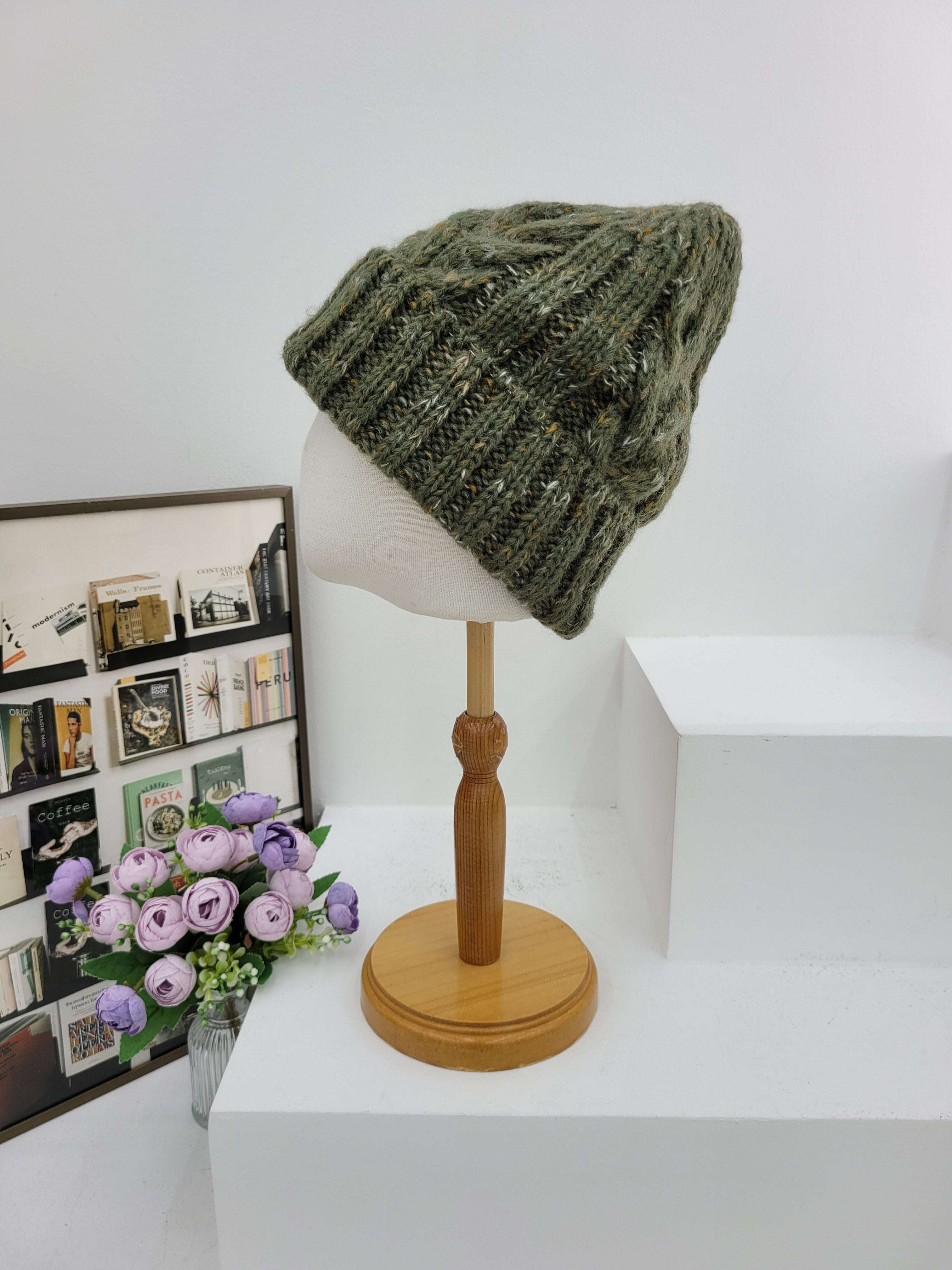 Green knitted hat on a mannequin head next to a wooden lamp and framed photos with flowers.