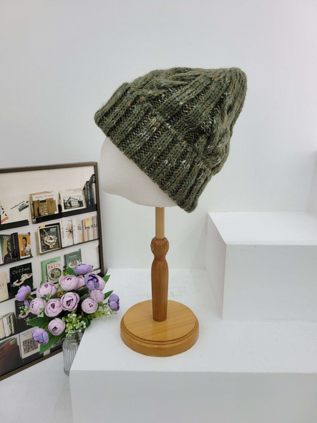 Green knitted hat on a mannequin head next to a wooden lamp and framed photos with flowers.