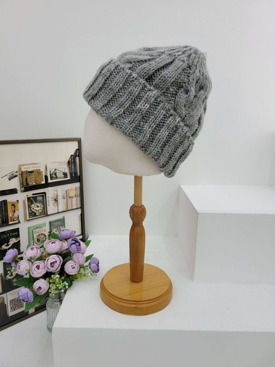 Gray knitted beanie on a mannequin head with a wooden lamp and floral arrangement in the background.