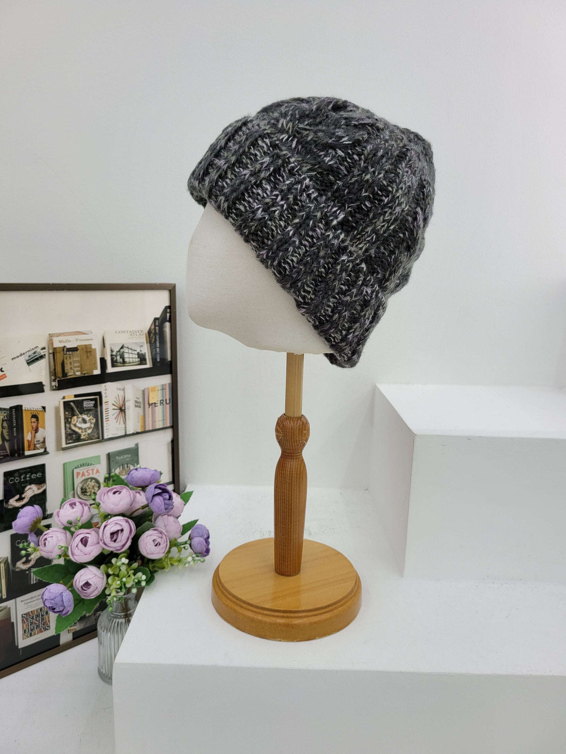 Gray knit beanie on a mannequin head against a white wall with a wooden lamp and floral arrangement.