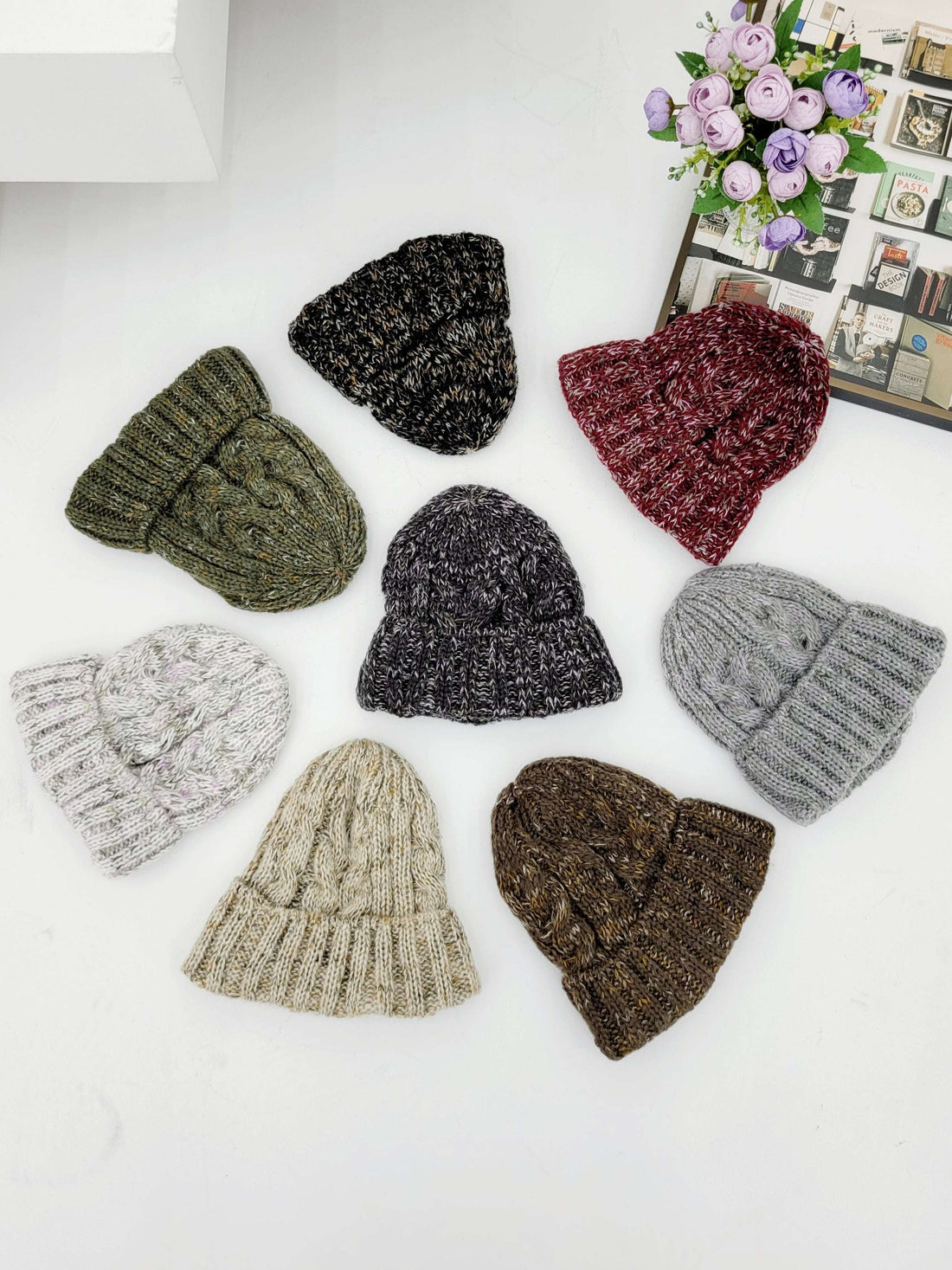 Nine knit hats in various colors on a white surface with a decorative background.