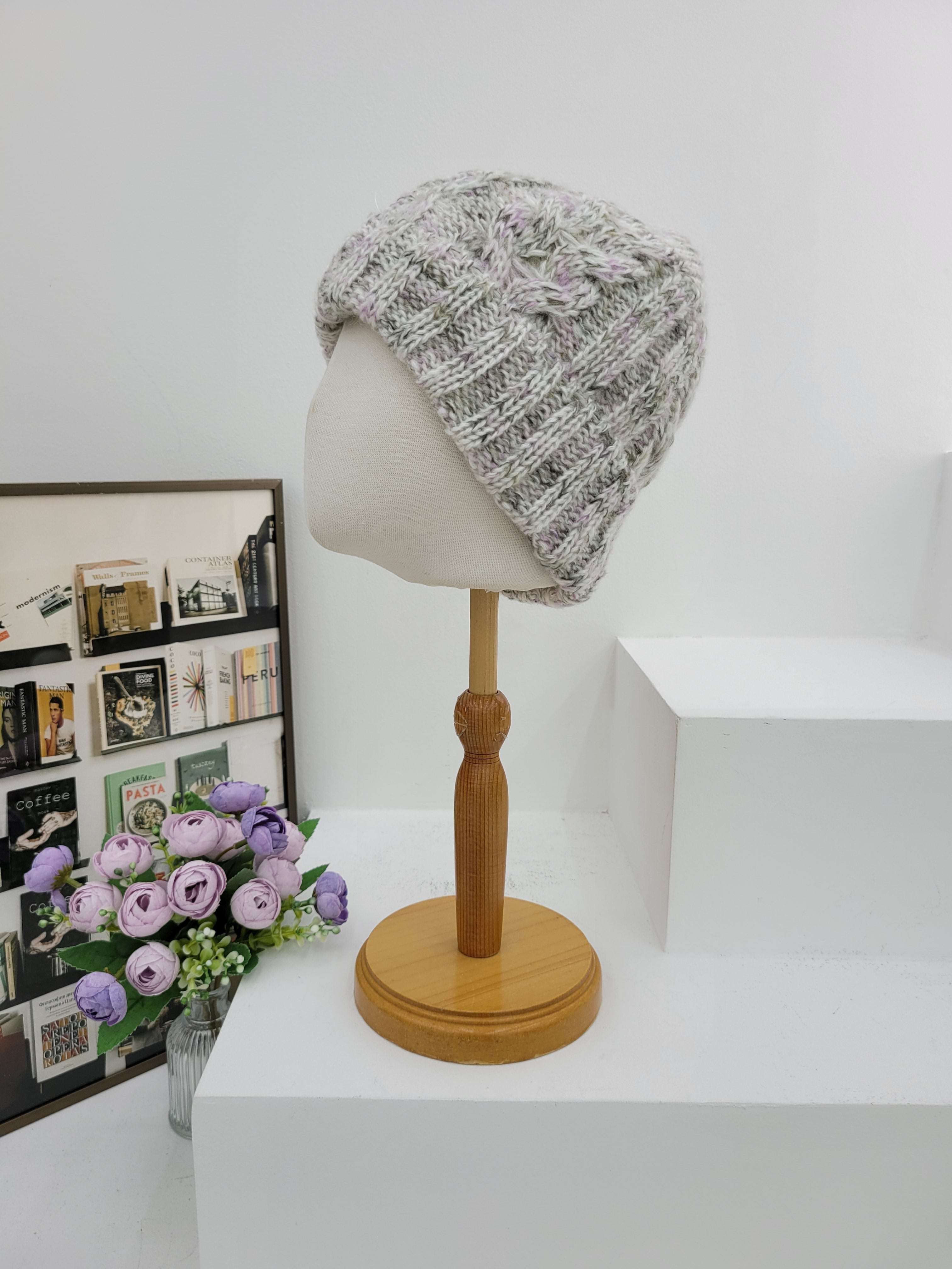 Knitted hat on a mannequin head next to a wall lamp with a wooden handle, on a white surface with a decorative box and flowers.