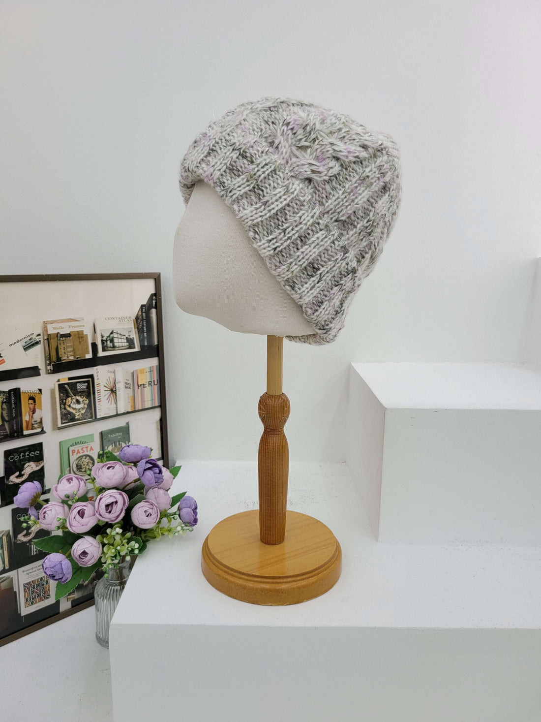 Knitted hat on a mannequin head next to a wall lamp with a wooden handle, on a white surface with a decorative box and flowers.