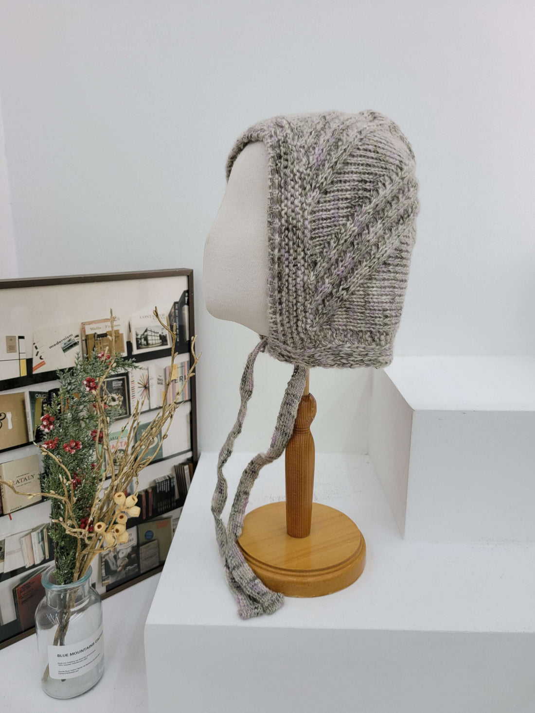 Knitted hat on a mannequin head against a white wall with a wooden rod and decorative items below.