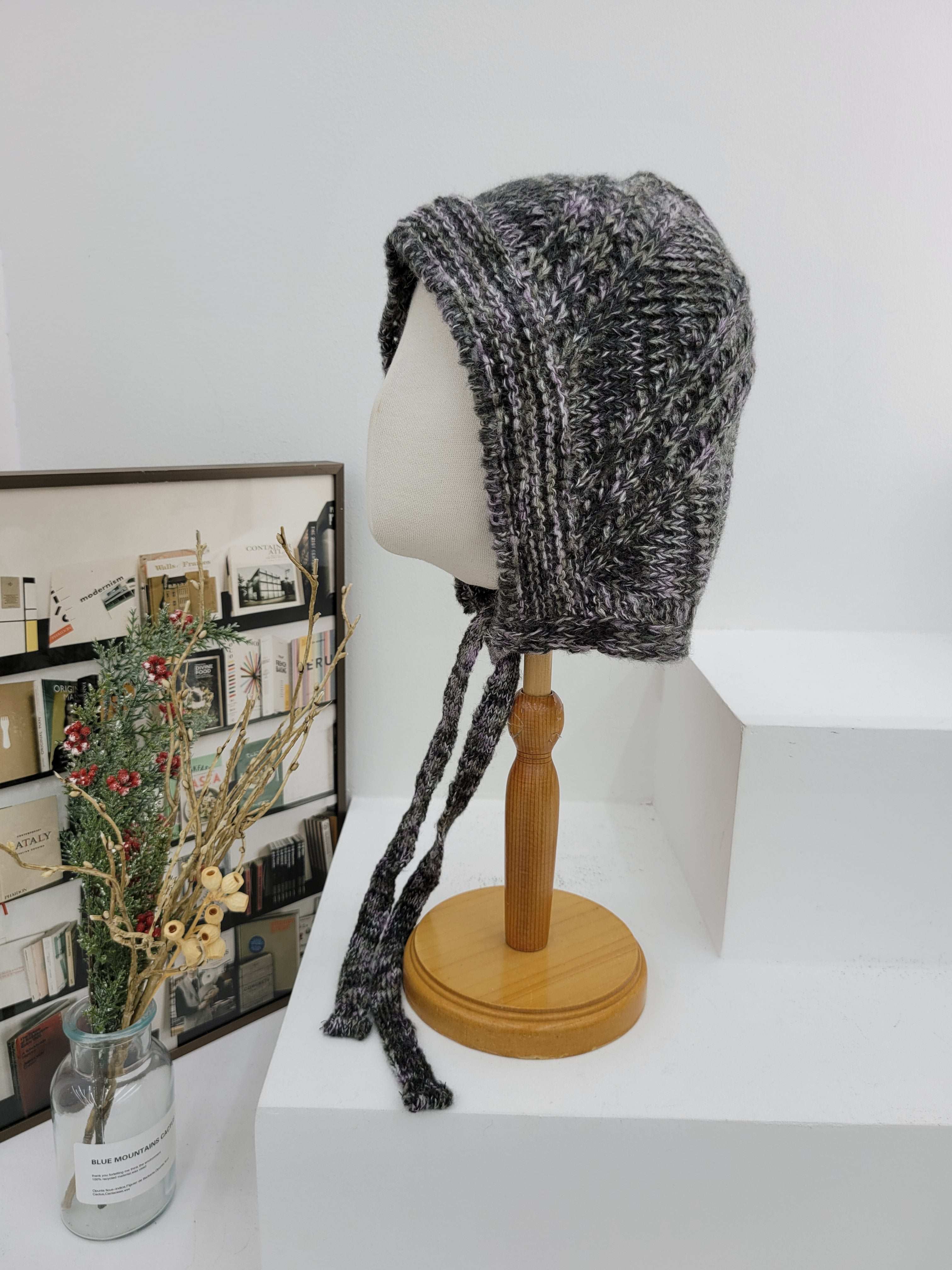 Knitted beanie on a wooden rod against a white wall with a decorative plant in the background.
