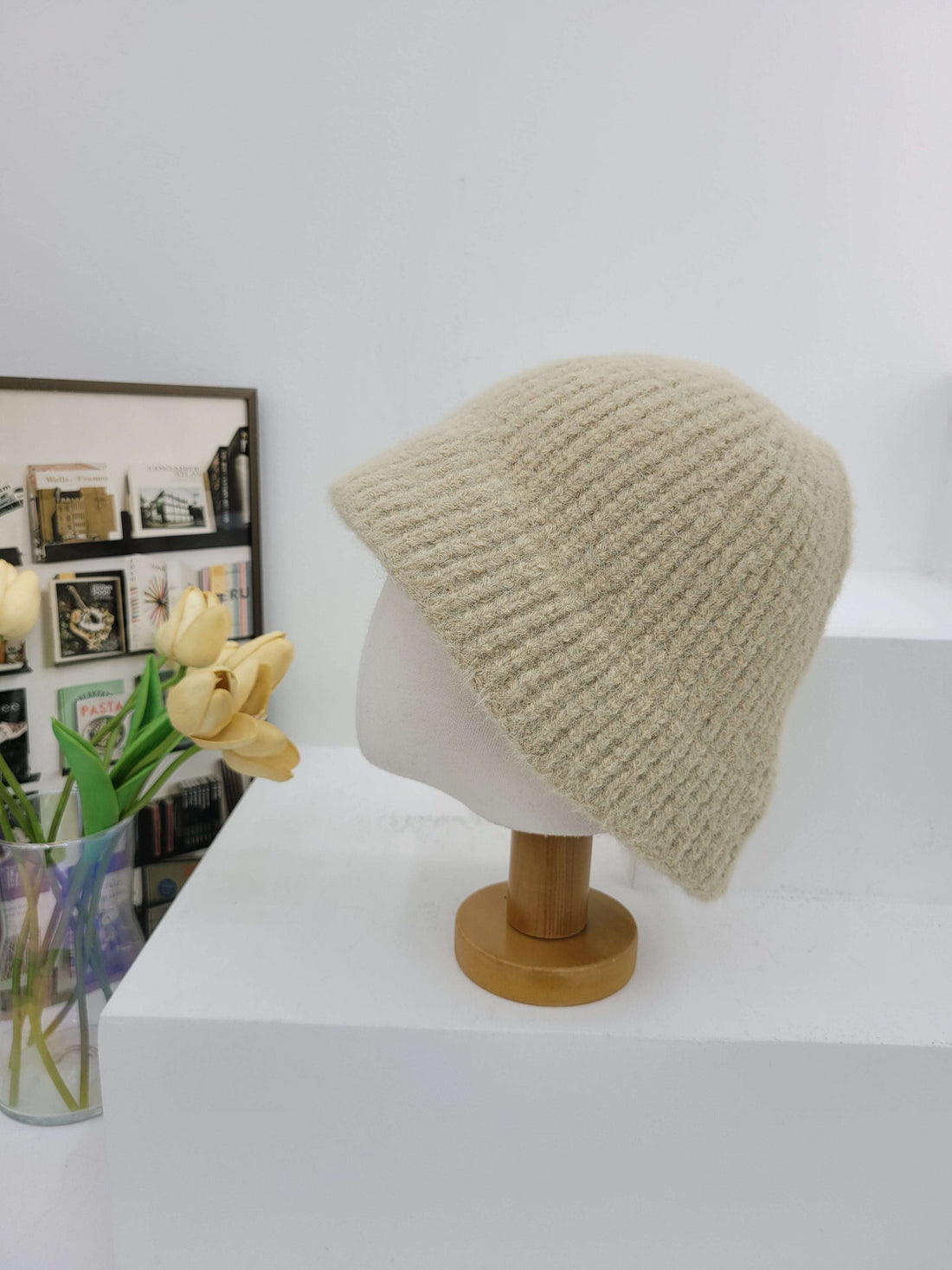 Knitted beige beanie on a mannequin head against a white wall with a vase of flowers below.