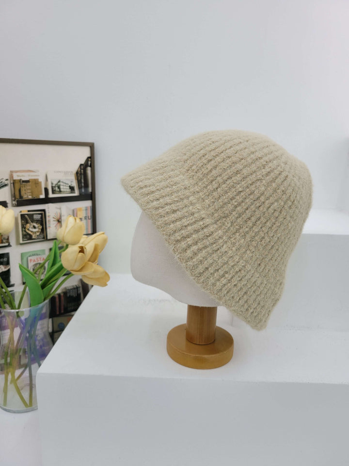 Knitted beige beanie on a mannequin head against a white wall with a vase of flowers below.