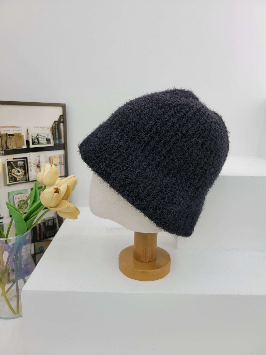 Black knit beanie on a mannequin head against a white wall with a decorative frame and flowers.