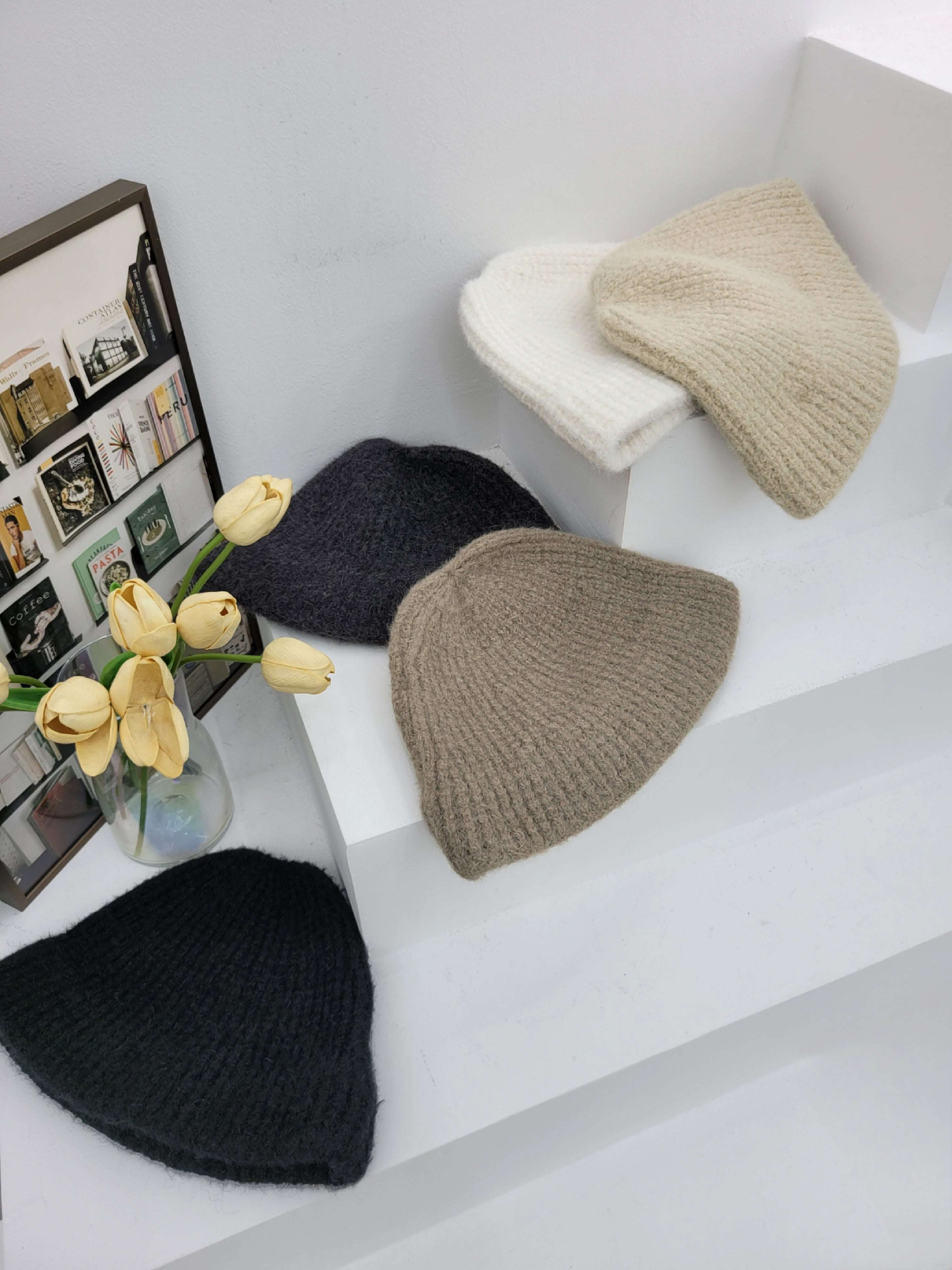 five knit hats in black, gray, and beige on a white surface with a vase of yellow tulips.