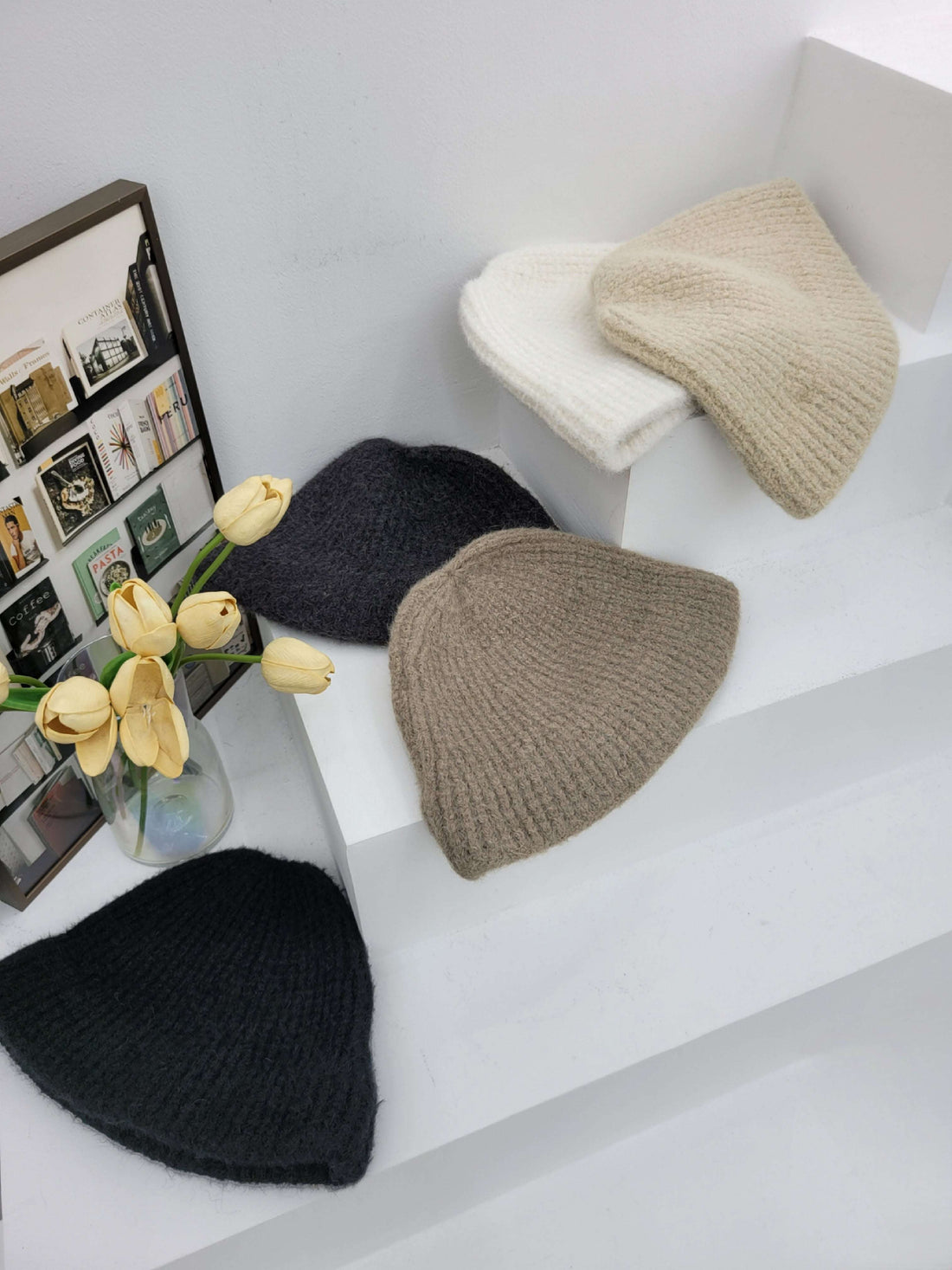 five knit hats in black, gray, and beige on a white surface with a vase of yellow tulips.
