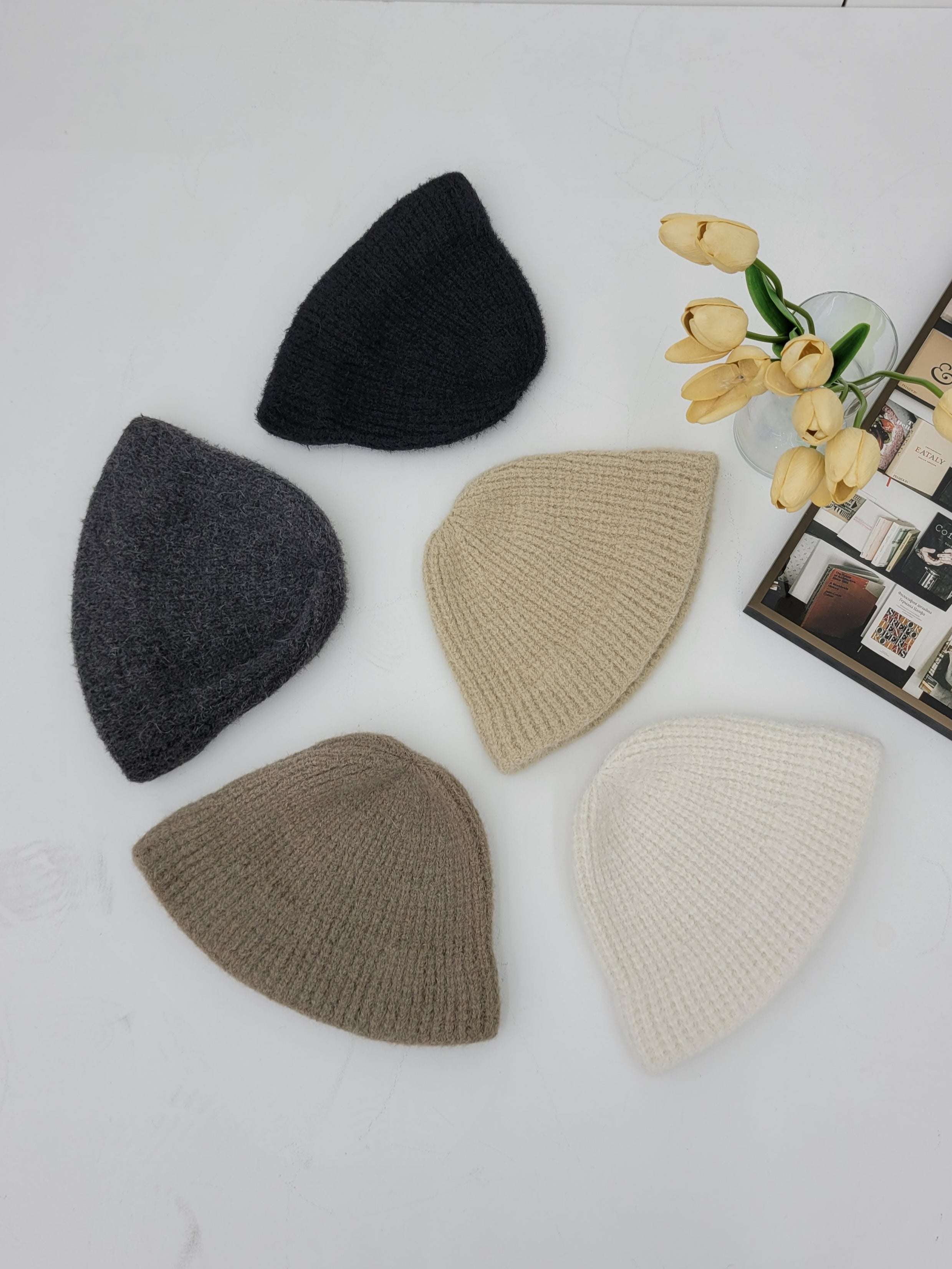 Five knit hats in black, gray, beige, and white on a light surface with yellow tulips.