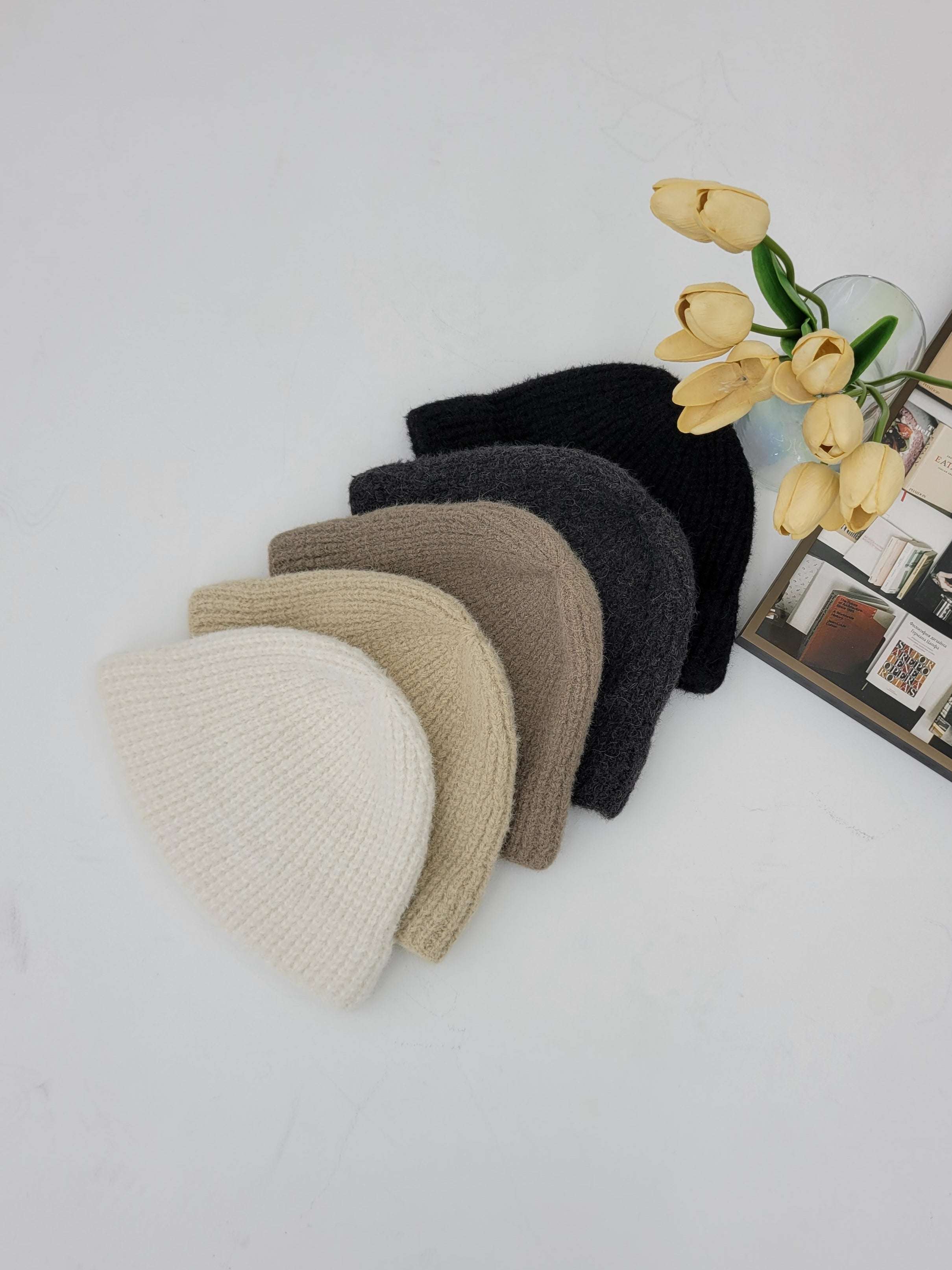 Set of knit beanies in various colors on a light surface with flowers and a photo frame in the background.