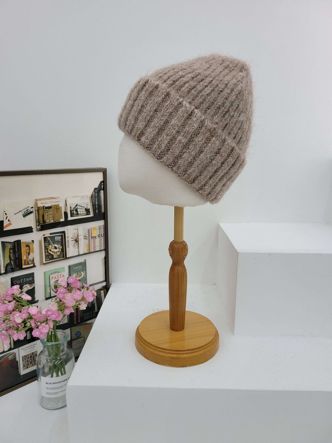 Beige knitted beanie on a mannequin head with wooden lamp against a white wall.