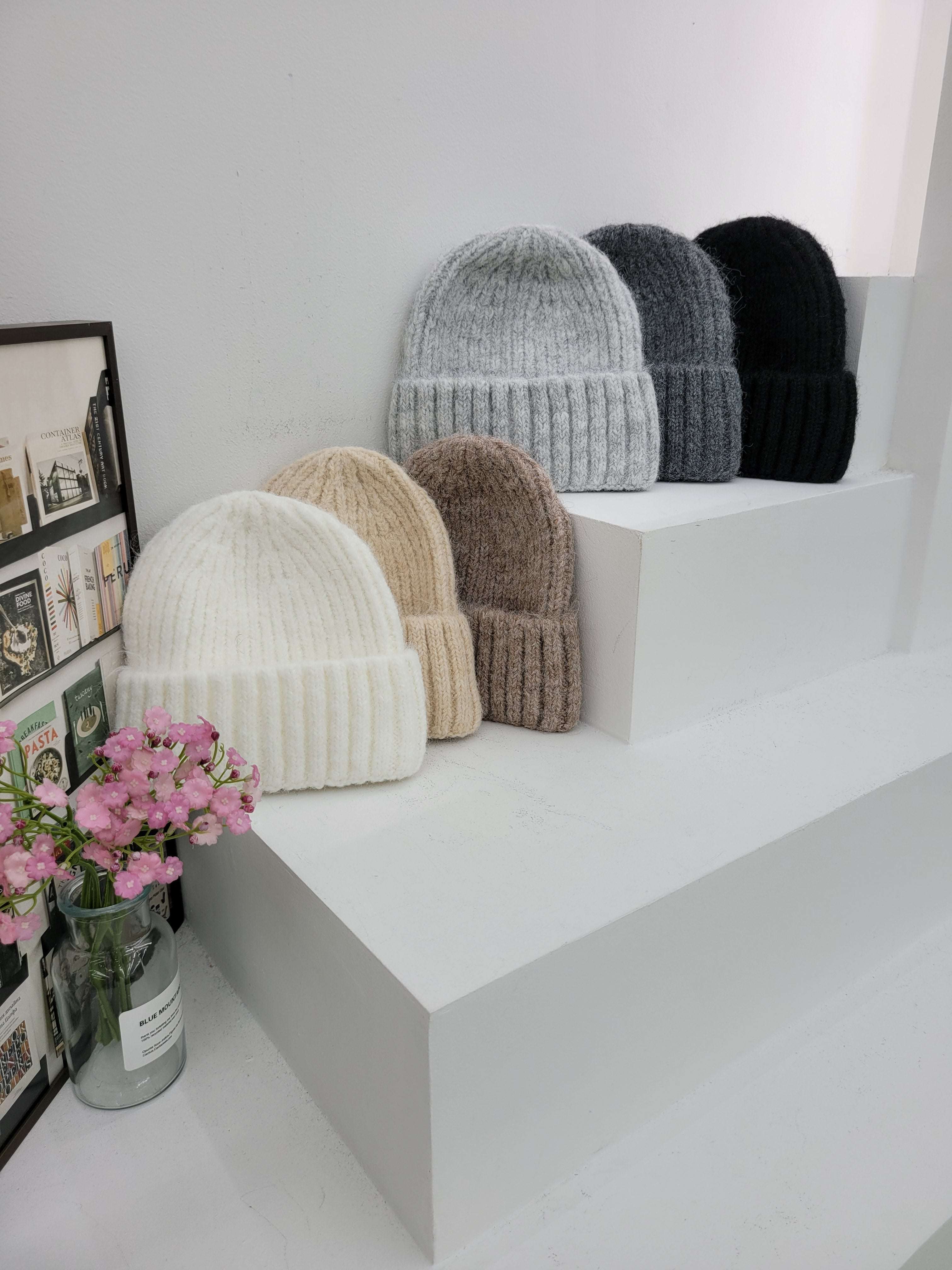 Stack of beanies cap in various colors on a white surface with a vase of pink flowers.