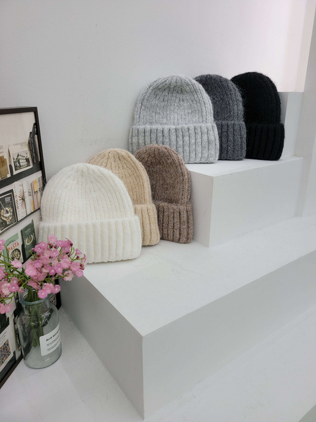 Stack of beanies cap in various colors on a white surface with a vase of pink flowers.