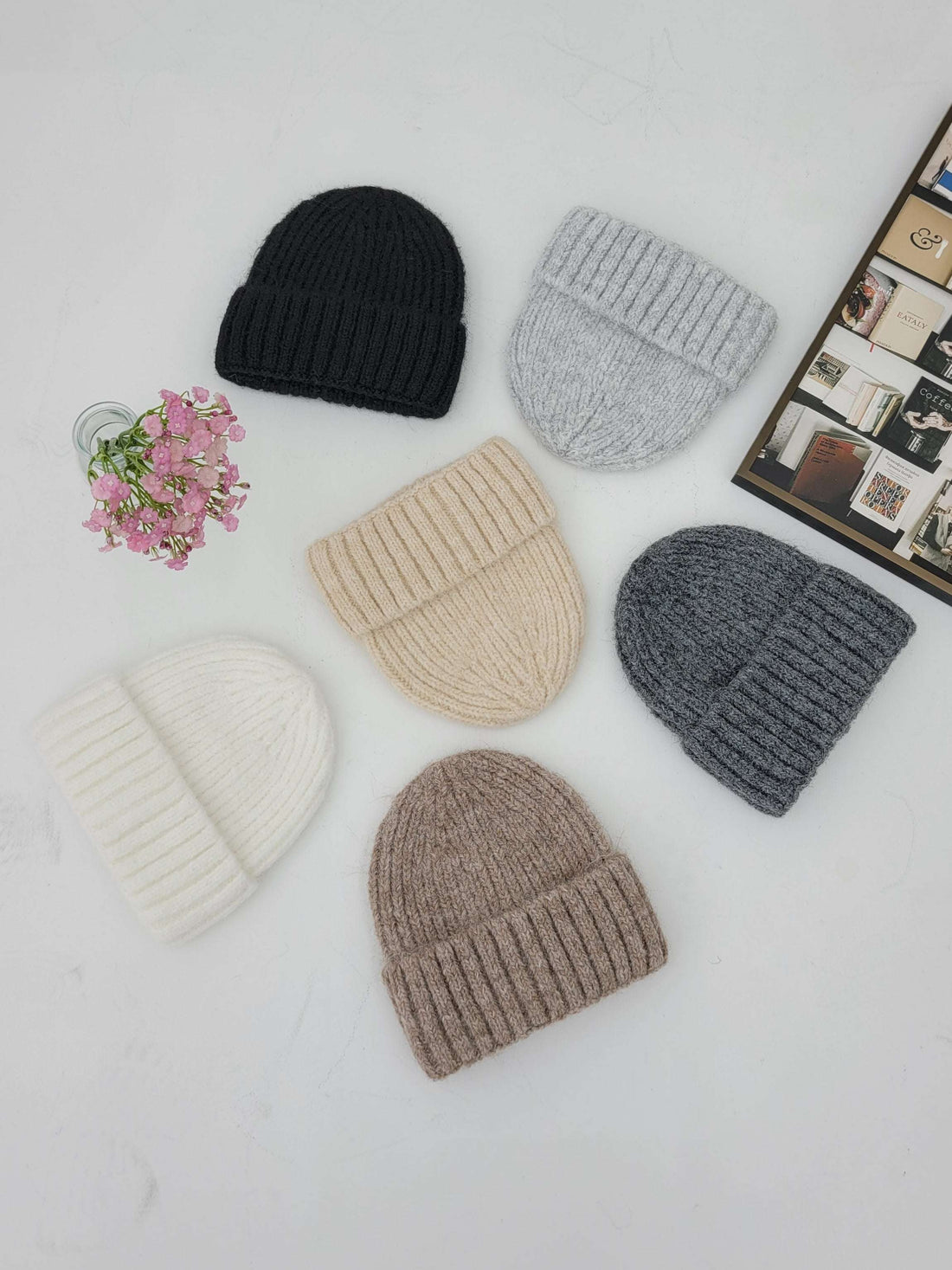 Six knit hats in black, gray, beige, and brown on a light surface with pink flowers.