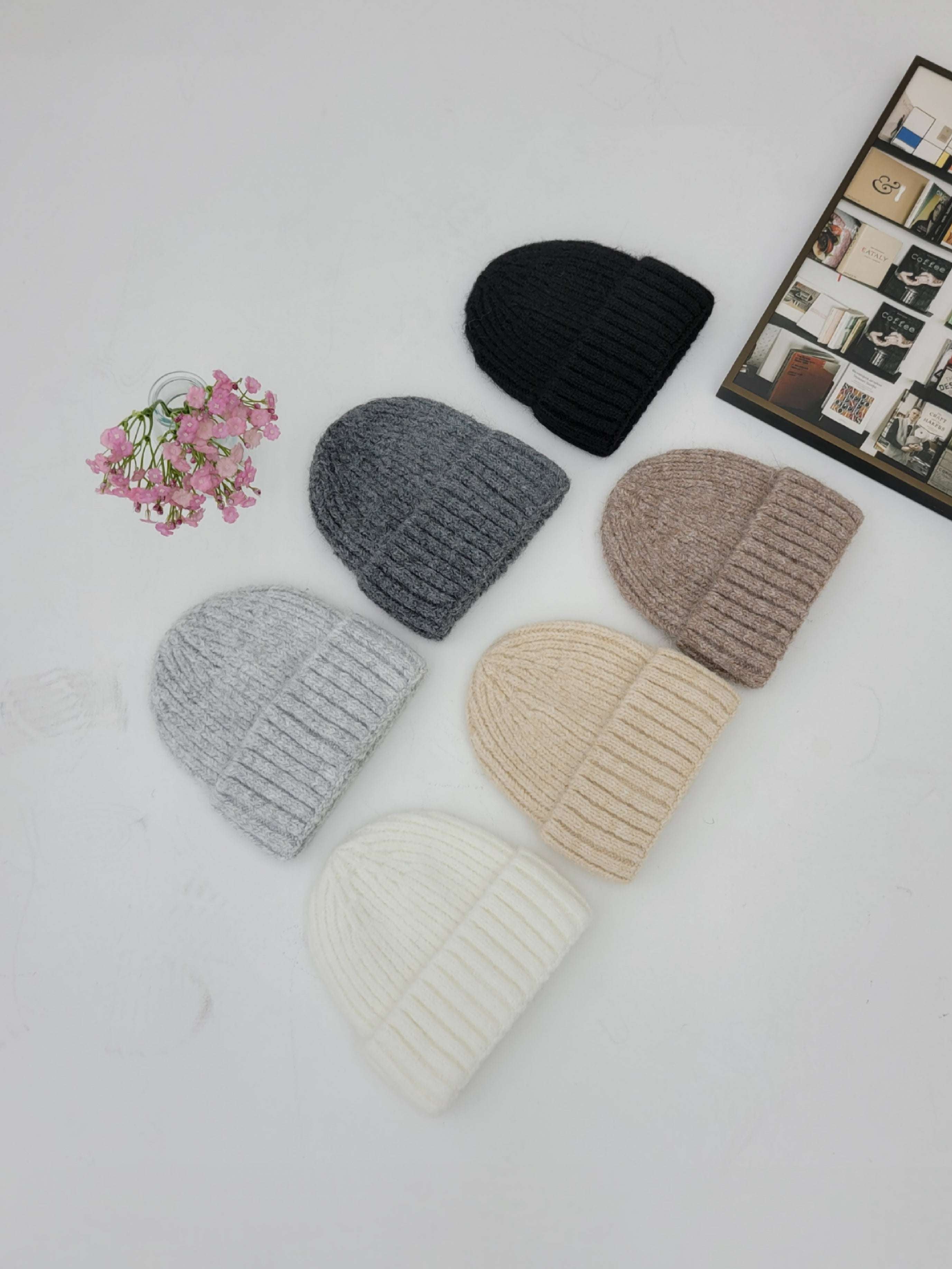 Five knit beanie hats in different colors on a white surface with a small bouquet of flowers and a framed picture.