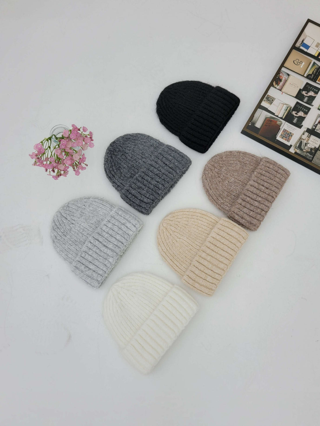 Five knit beanie hats in different colors on a white surface with a small bouquet of flowers and a framed picture.