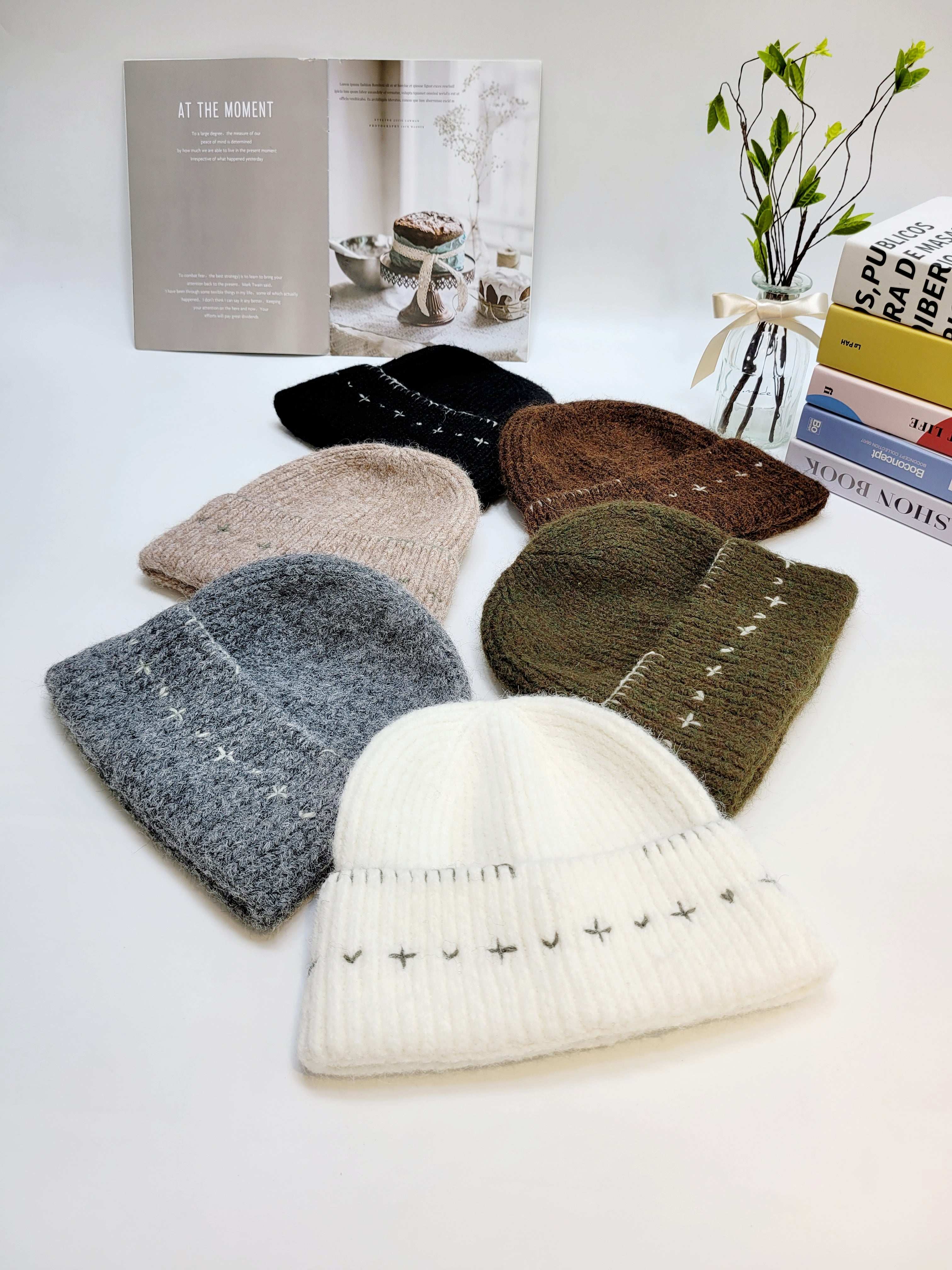 Collection of knit hats in various colors on a white surface with books and a plant in the background.