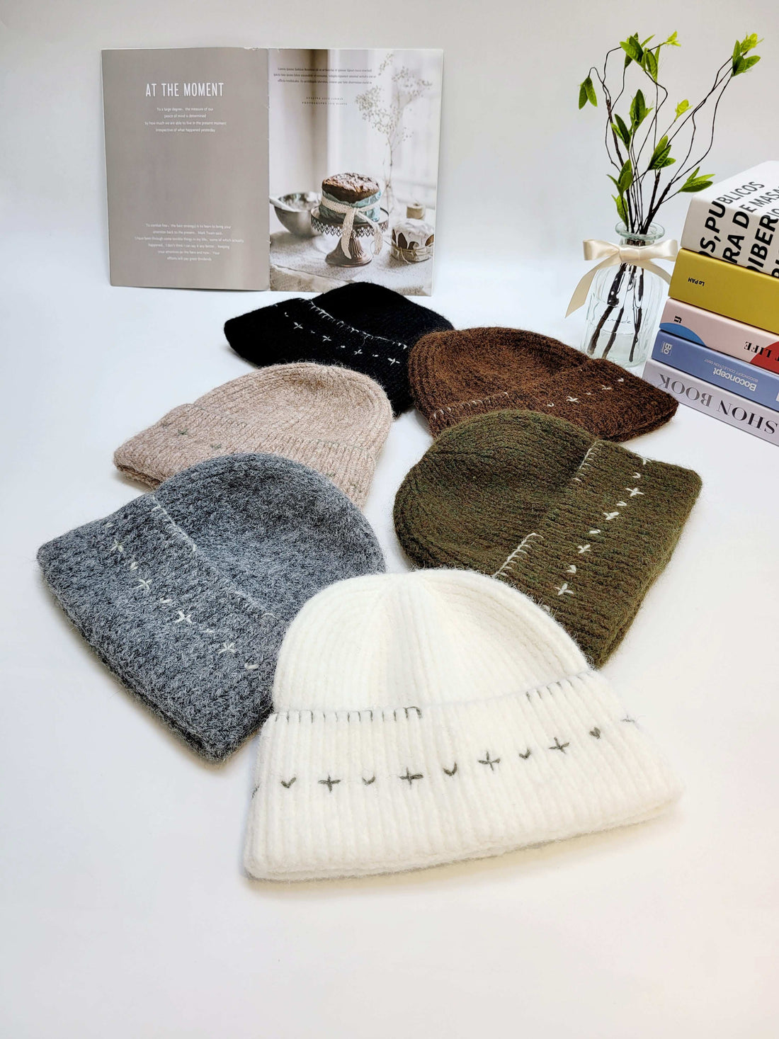 Collection of knit hats in various colors on a white surface with books and a plant in the background.