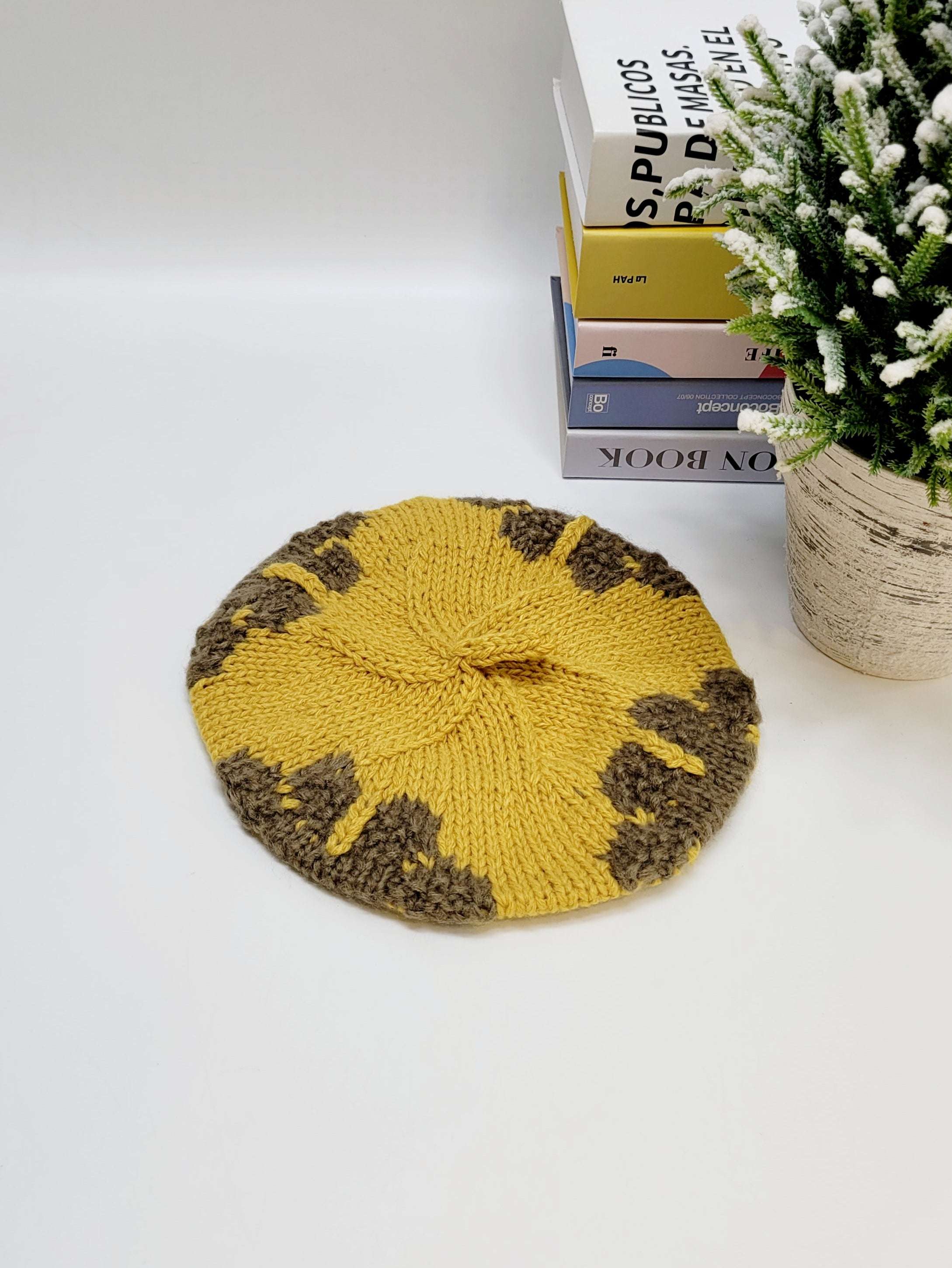 Yellow knitted beret on a white surface with books and a plant in the background