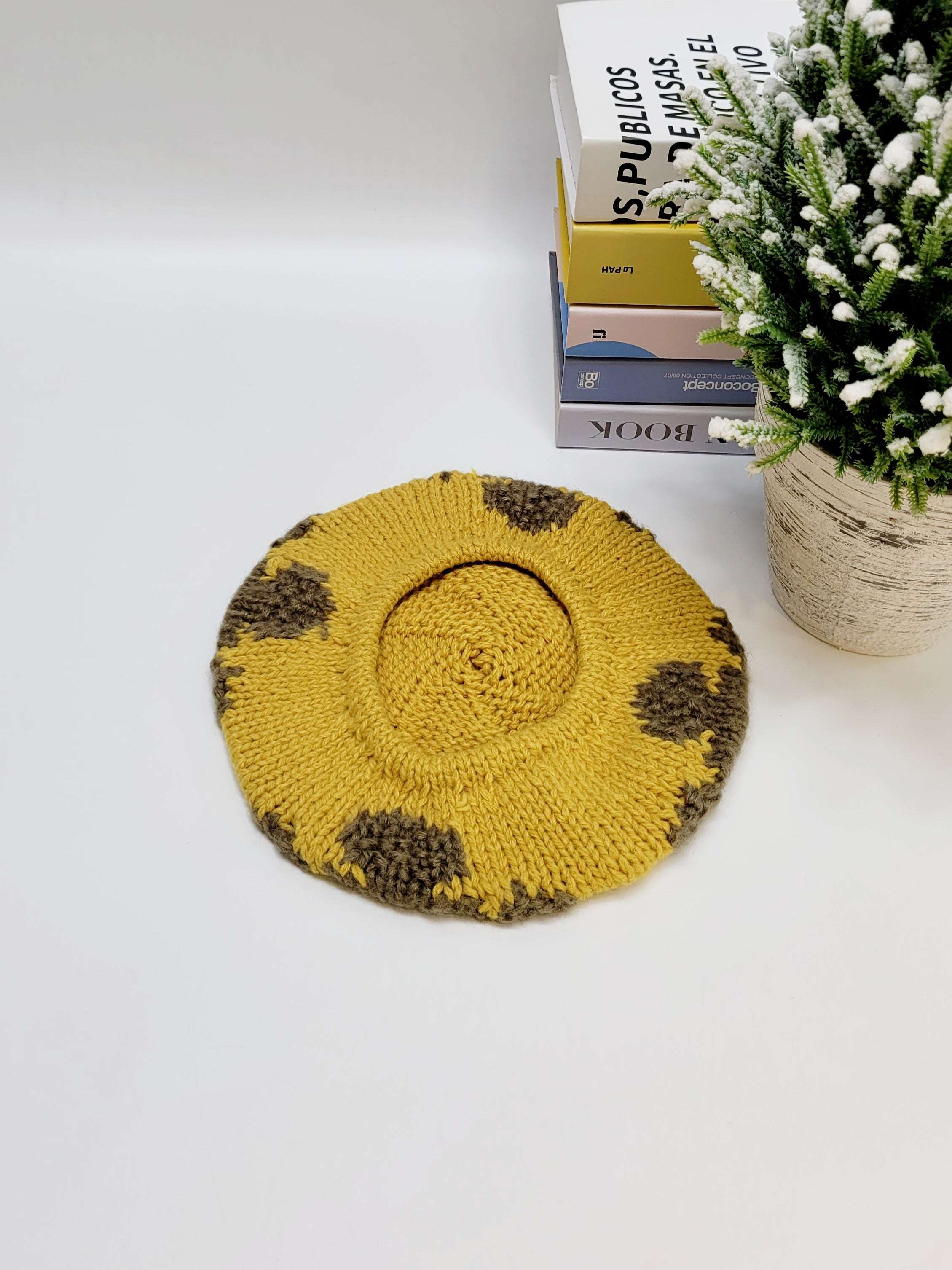 Yellow knitted beret with brown spots on a white surface with books and a plant in the background.