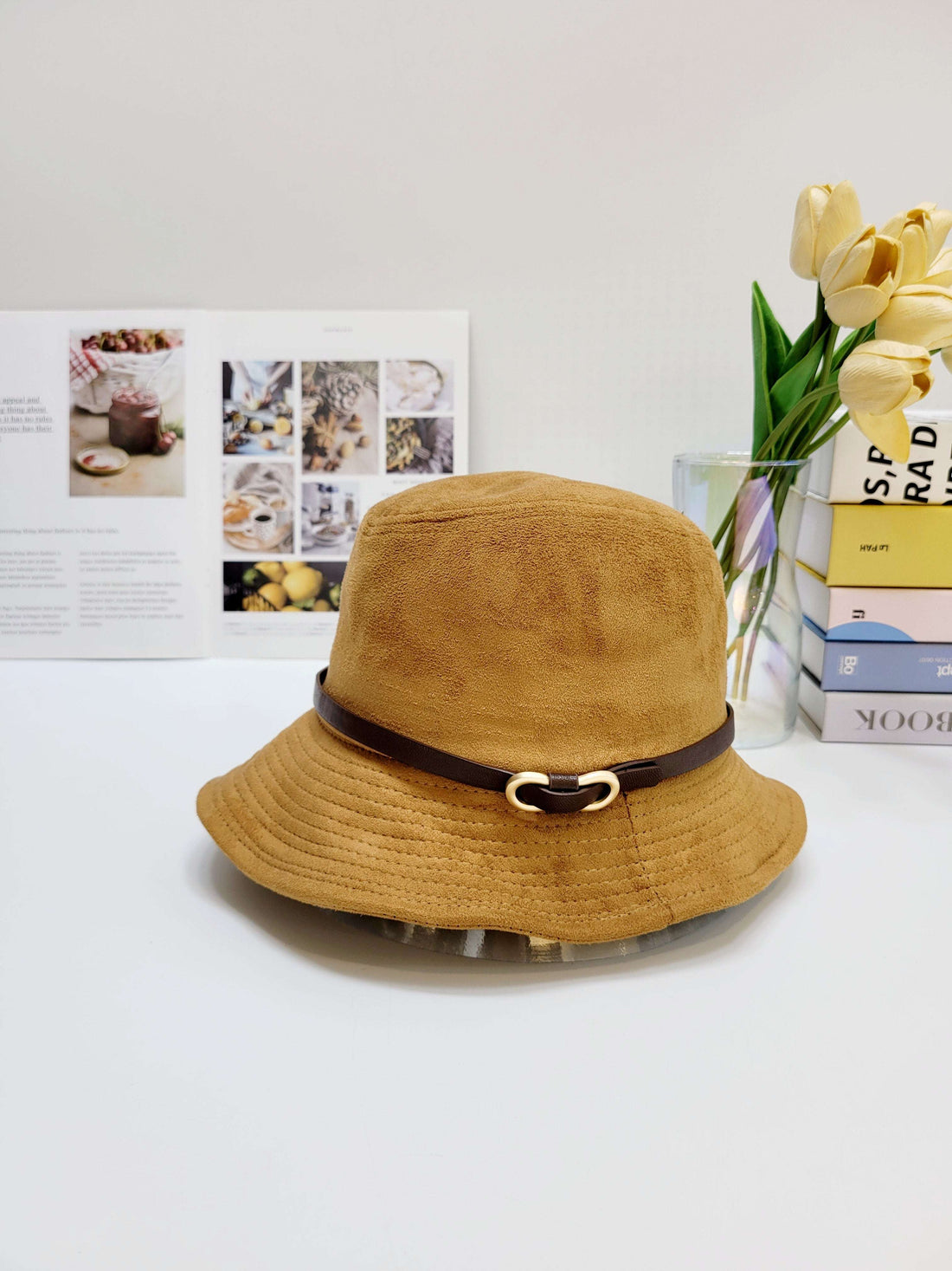 Brown bucket hat with a belt on a white surface with a magazine and flowers in the background