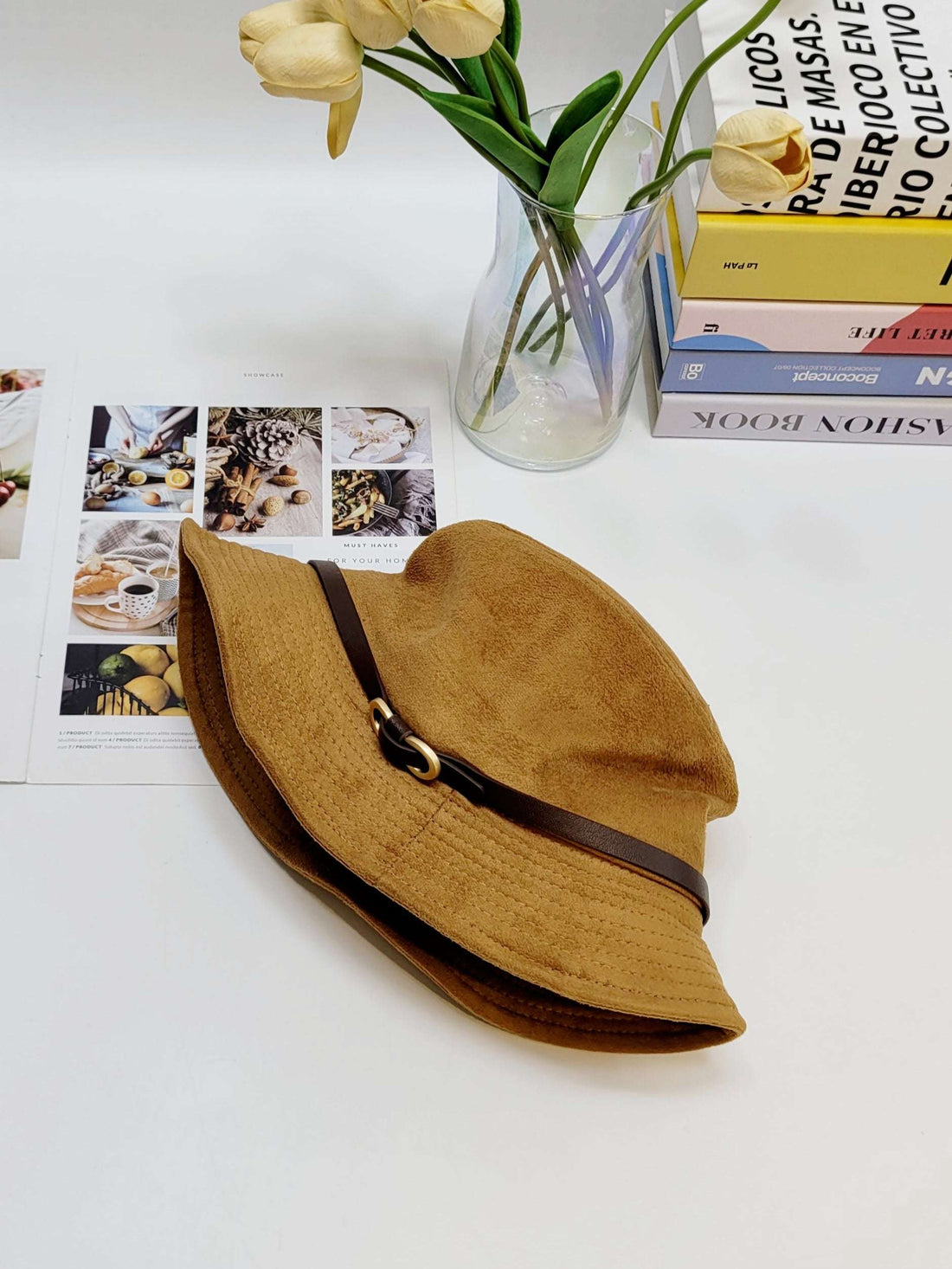 Brown hat with a brown ribbon on a white surface with a vase of flowers and books in the background.