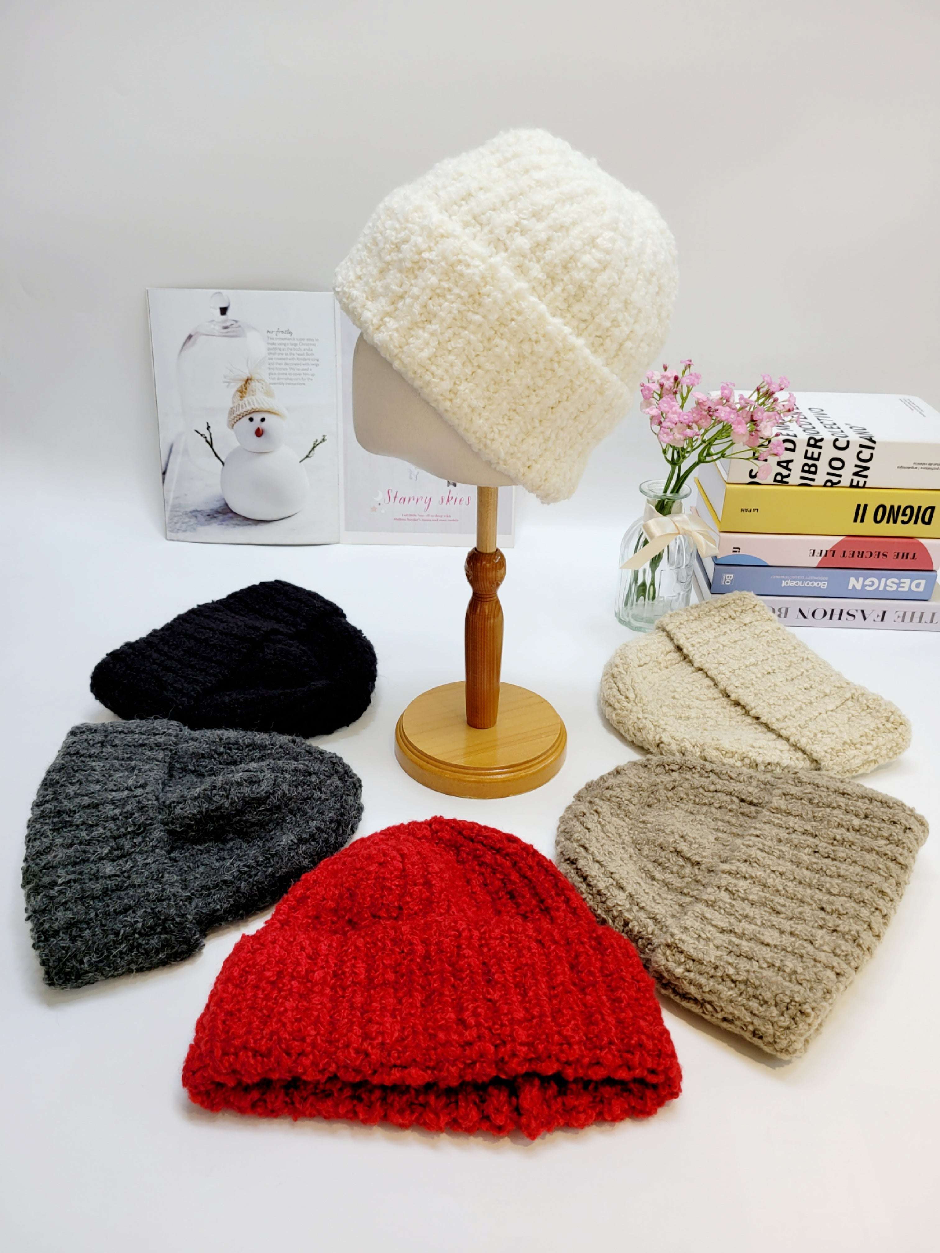 Set of knitted hats in various colors on a white surface with a small plant and books in the background.