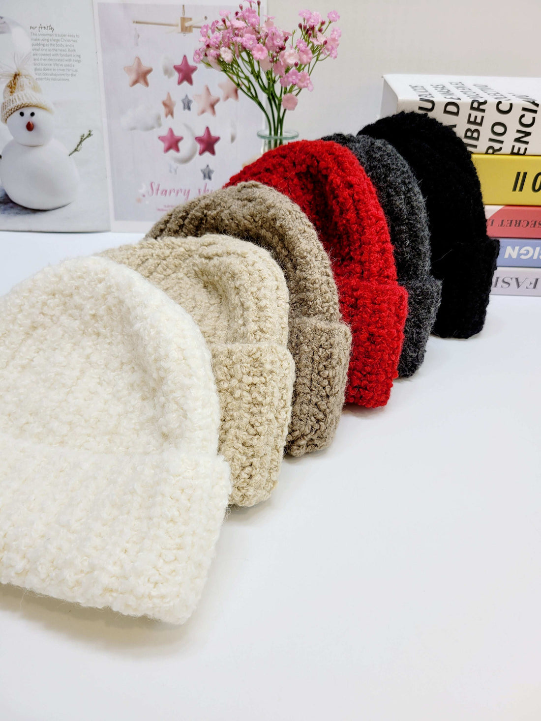 Set of colorful knitted beanies on a white surface with books and flowers in the background.