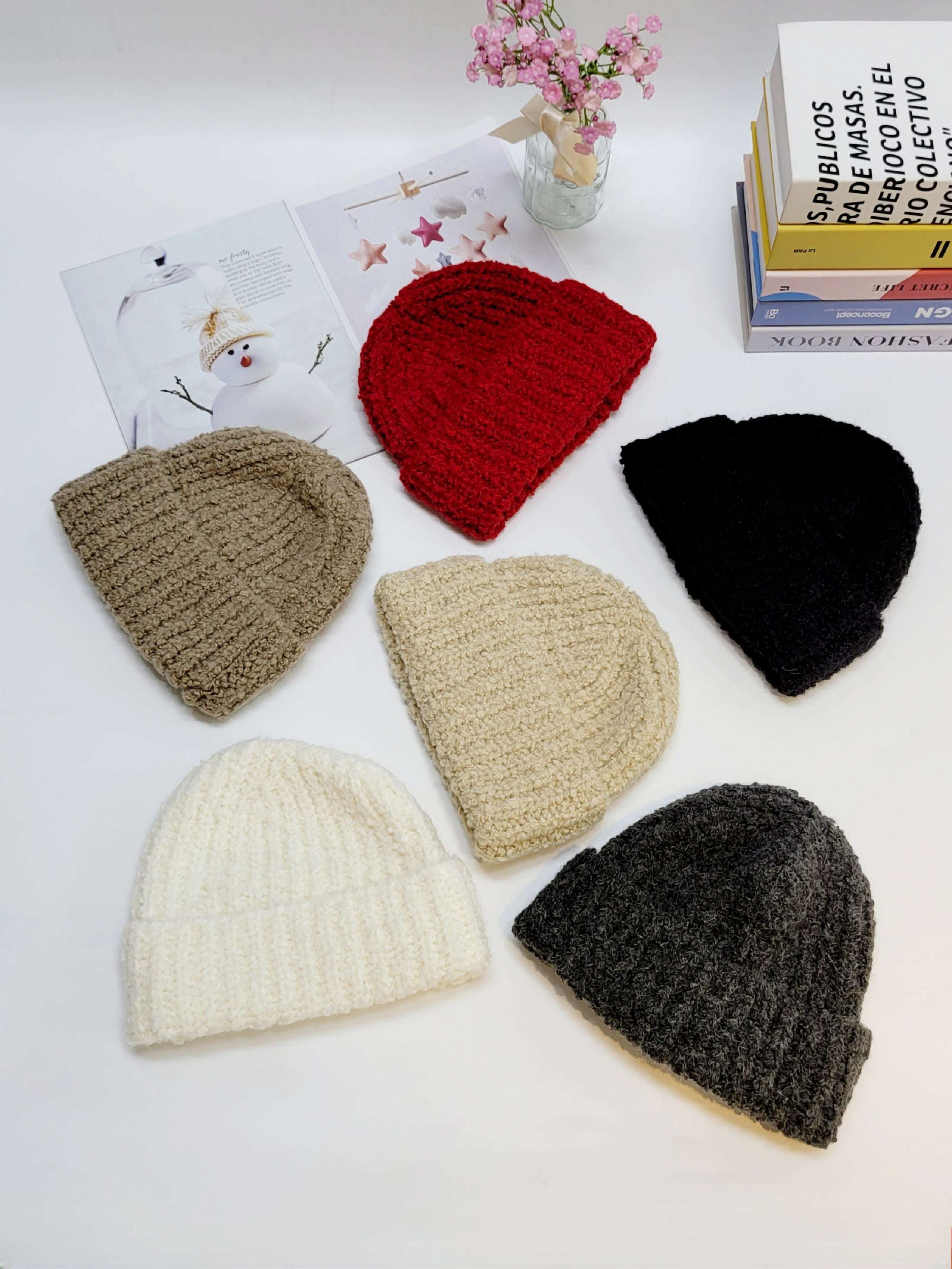 Set of six knitted hats in various colors on a white surface with books and flowers in the background.