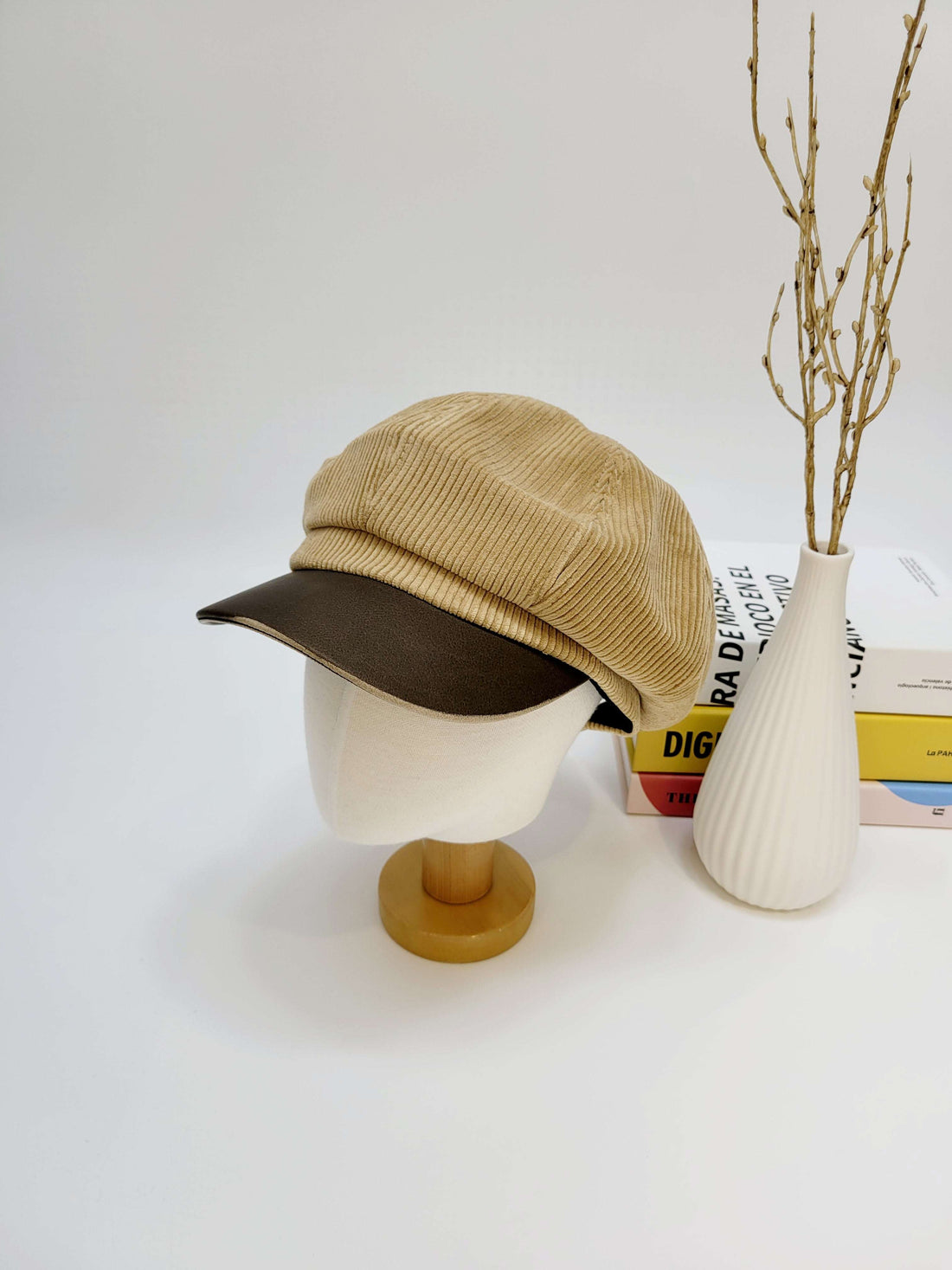 Beige cap on a mannequin head with a white vase and books in the background