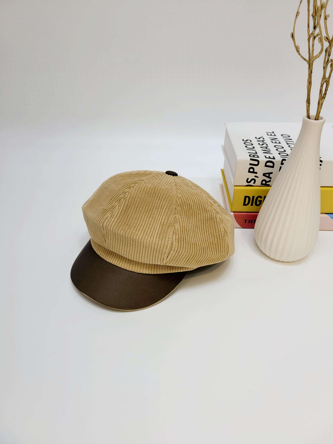 Beige corduroy cap with brown visor on a white surface with books and a vase in the background.