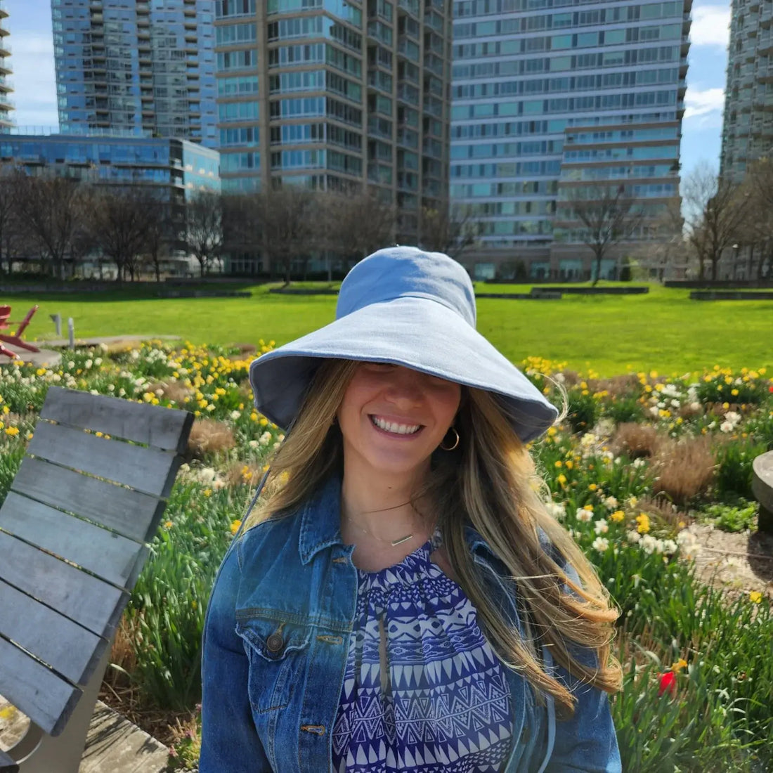 Woman smiling in a cotton linen wide brim-solid hat, light blue, with denim jacket, among spring flowers.