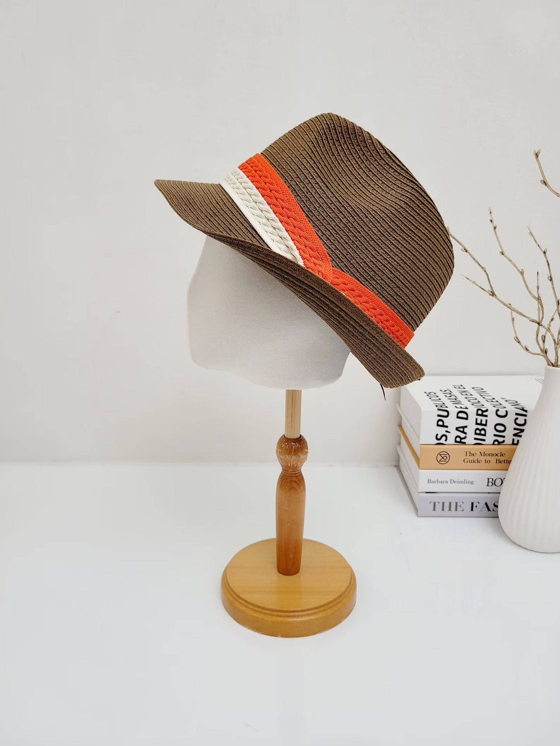 Brown raffia fedora hat with a stylish orange, white, and beige two-tone band, displayed on a wooden stand.